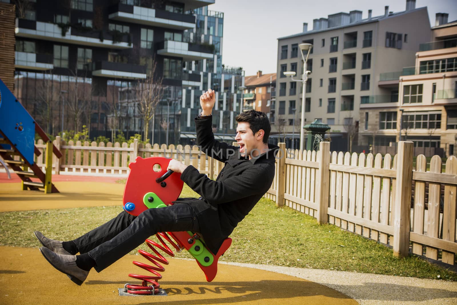 Young man reliving his childhood plying in a children's playground riding on a colorful red spring seat with a happy smile in an urban park