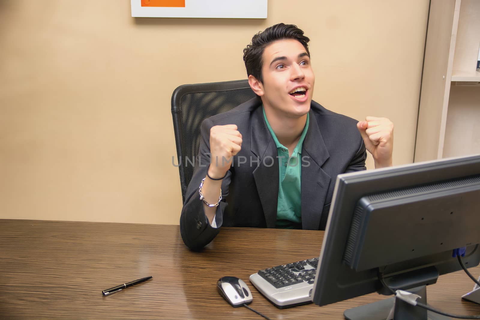 Happy young businessman screaming for joy, sitting at desk in his office working with computer