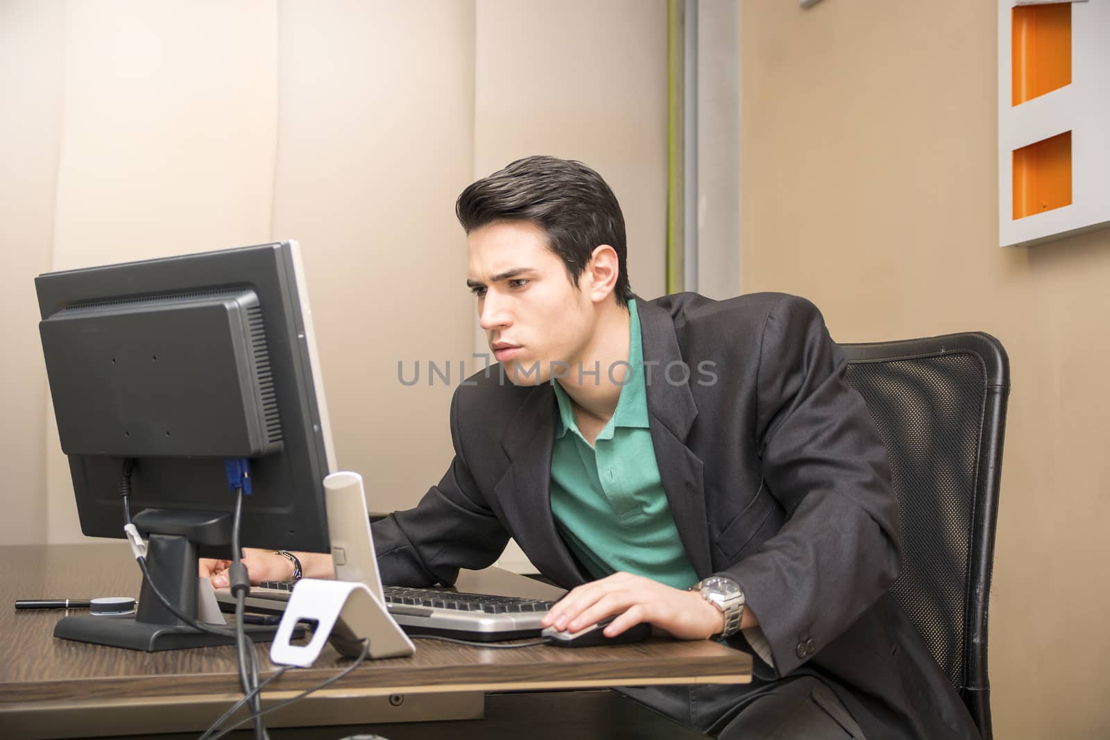 Serious handsome young businessman sitting at his desk in the office working at computer