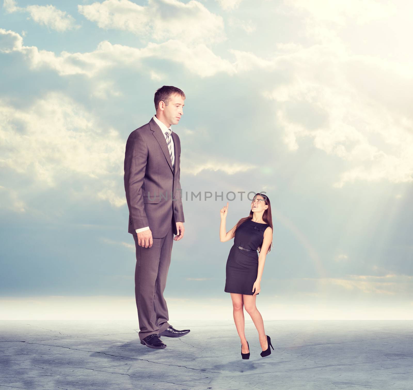 High businessman looking down at little woman in dress. Clouds and cement surface as background. Business concept