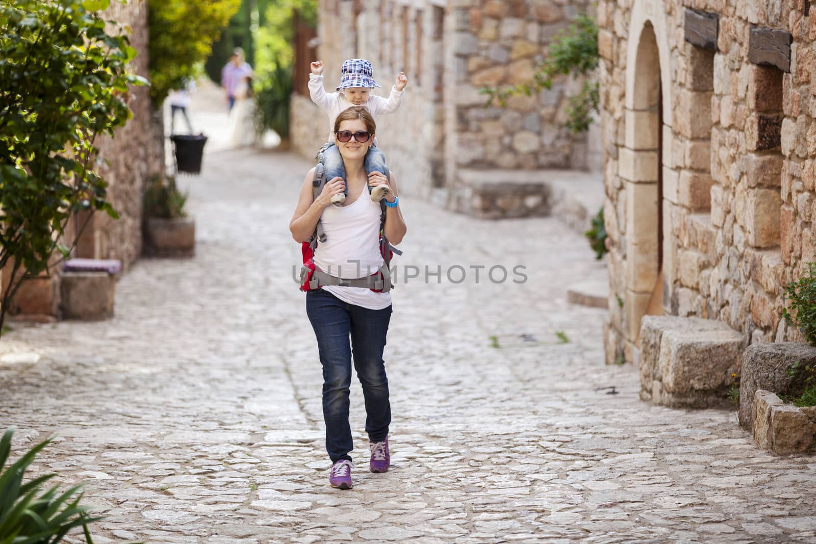 Young Caucasian woman tourist carrying her little son on shoulders while exploring old Spanish town