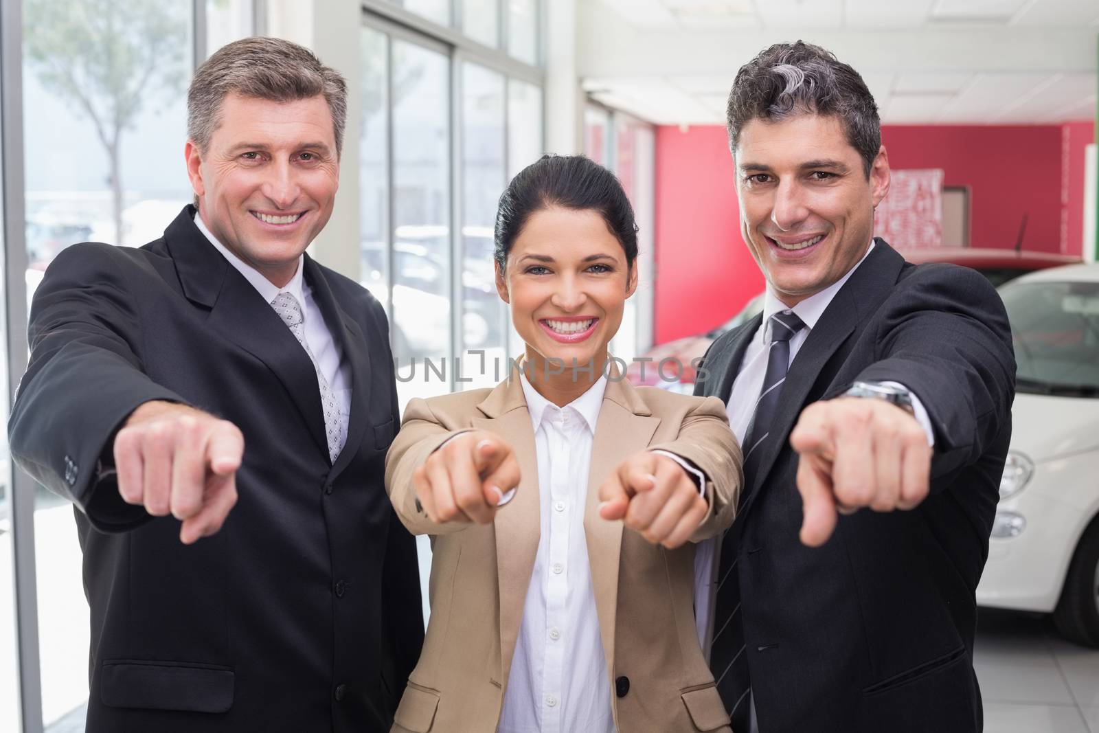 Smiling business team pointing at camera at new car showroom