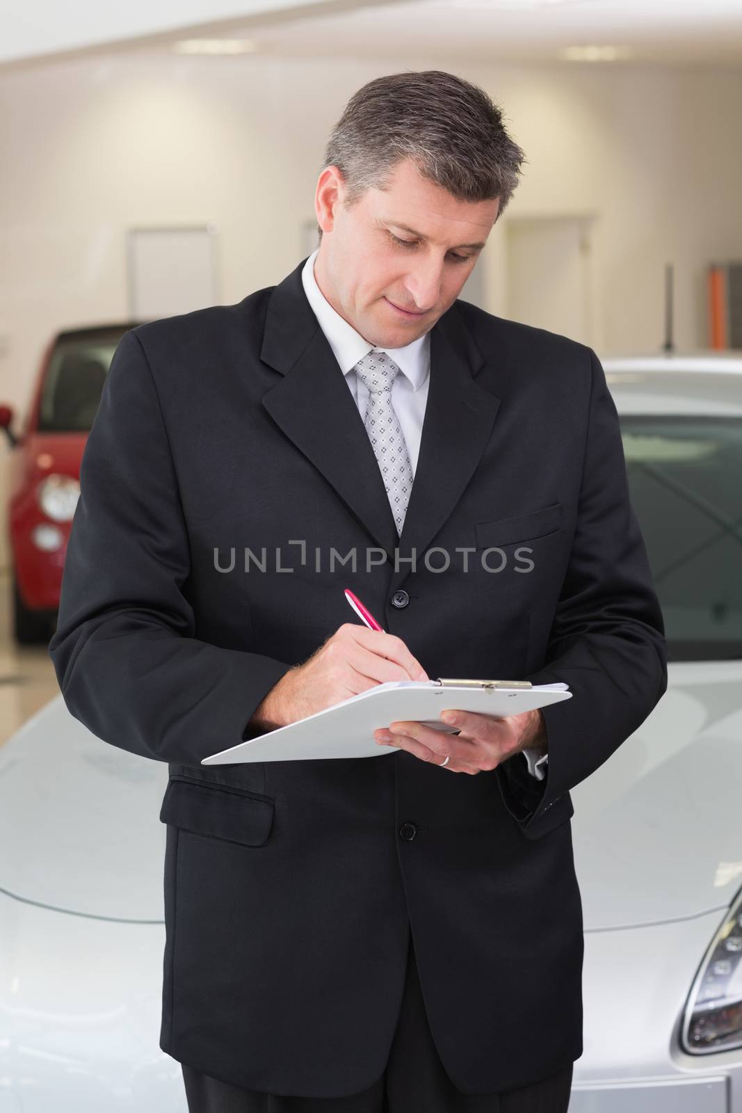 Serious businessman writing on clipboard at new car showroom