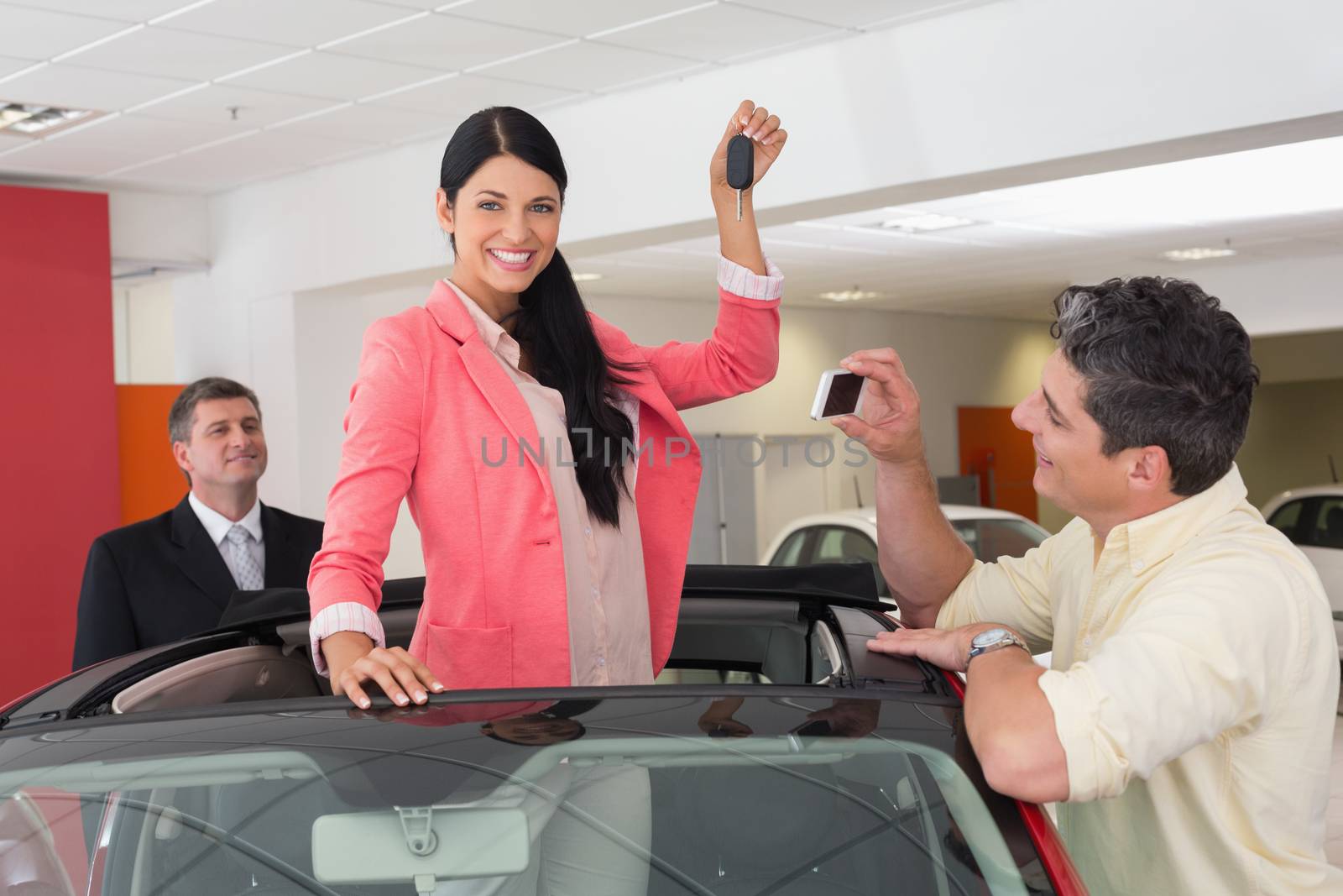 Woman standing in red cabriolet and man taking picture at new car showroom