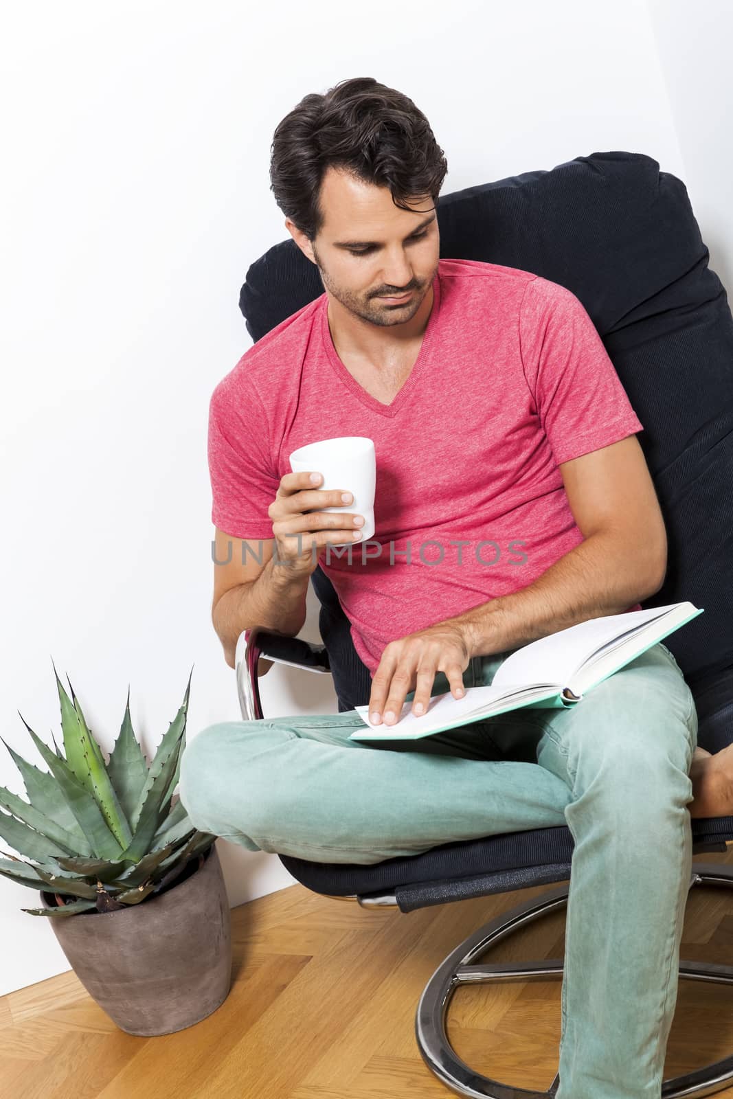 Young Man in Casual Clothing Sitting on Black Chair While Reading a Book and Holding a Glass of Drink.