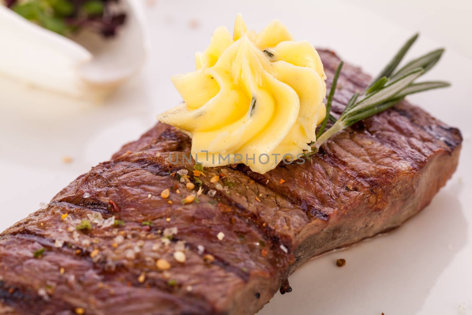 Tasty grilled beef steak topped with a twirled knob of butter and a sprig of fresh rosemary and served on a white plate, close up view