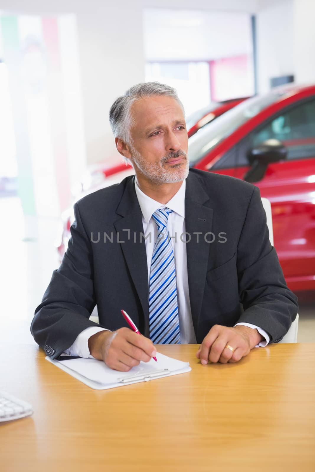 Focused salesman writing on clipboard at his desk at new car showroom