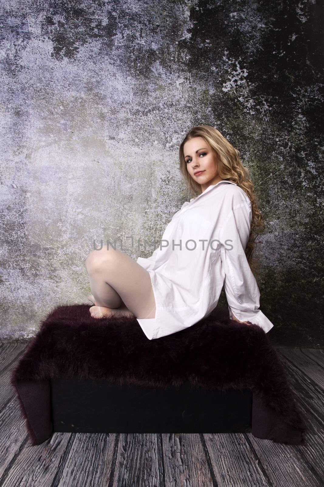 Young long-haired curly blonde woman on the sofa with a hairy blanket in front of an old dilapidated wall