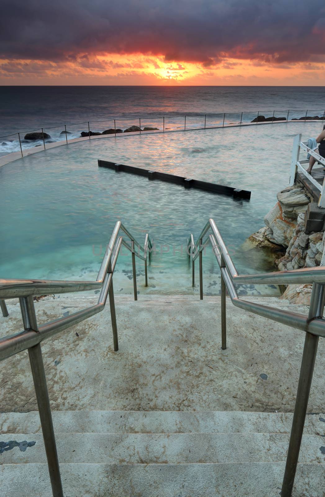 The steep steps leading down into Bronte Ocean Baths at the southern end of Bronte Beach, Sydney Australia.  The pool is built into the cliffs and has a unique kickboard for lap swimmers.The sun breaks free of clouds at sunrise casting a soft orange glow onto the clouds and across the ocean.