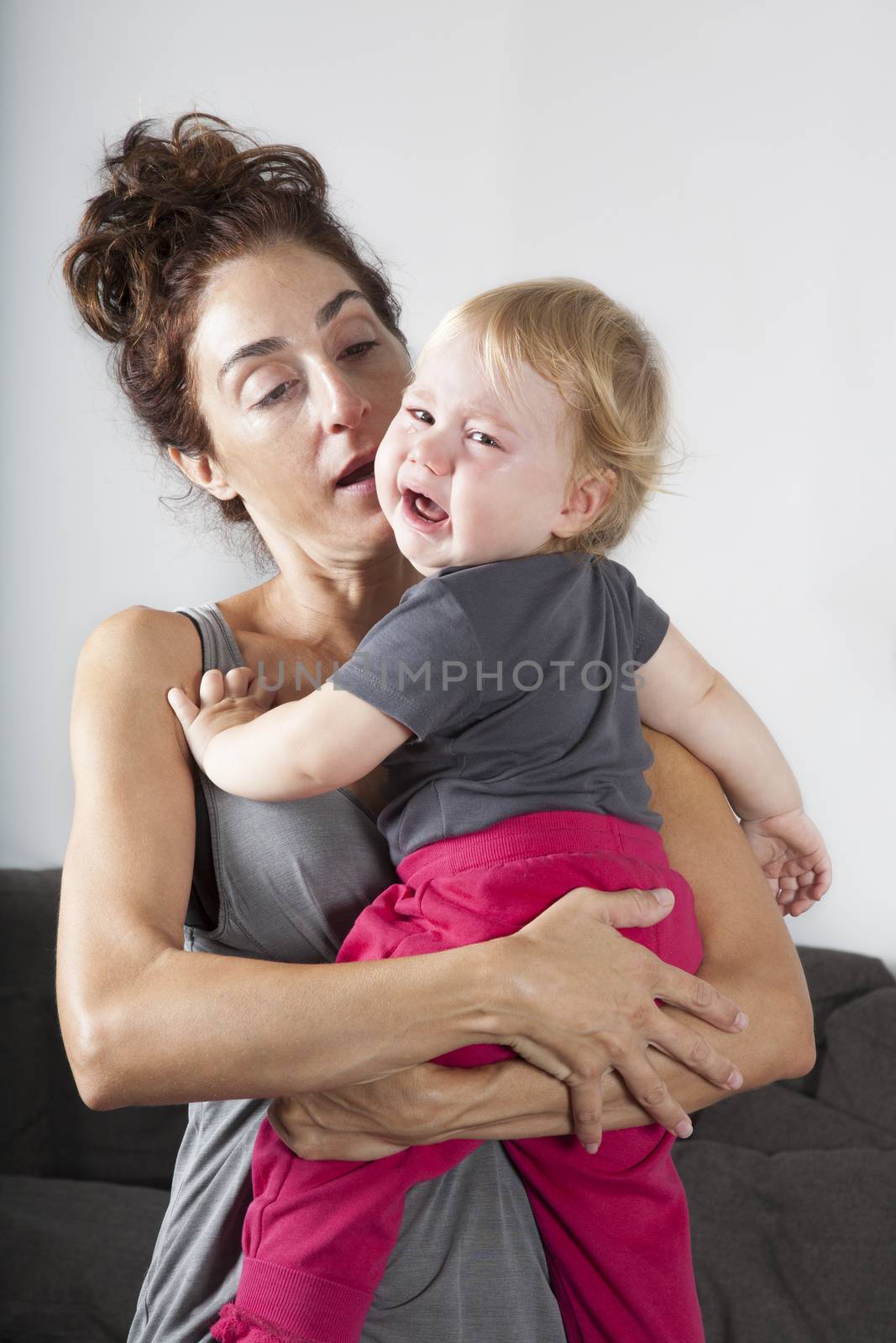 portrait of one year age blonde lovely cute caucasian white baby grey shirt pink trousers looking at camera face indoors crying and scream shout with tears eyes in brunette woman mother arms embrace hug