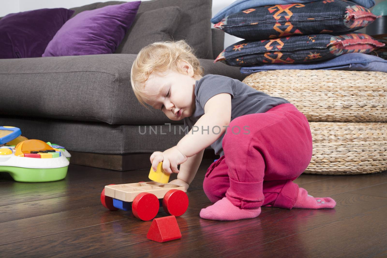 one year age blonde lovely cute caucasian white baby grey shirt pink trousers and shocks playing with wheel wooden colors toys indoor on brown floor