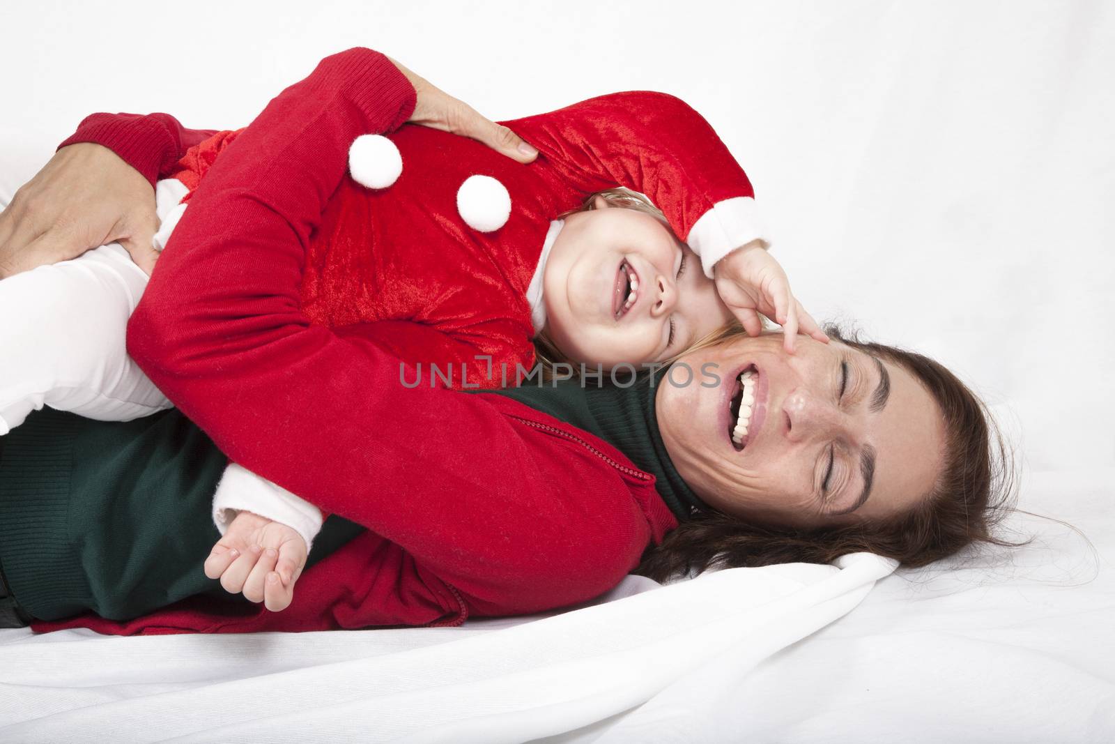 tender and funny portrait of one year age caucasian blonde cute lovely baby Santa Claus Christmas disguise with brunette woman mother red cardigan green sweater embraced laughing together lying on white floor background