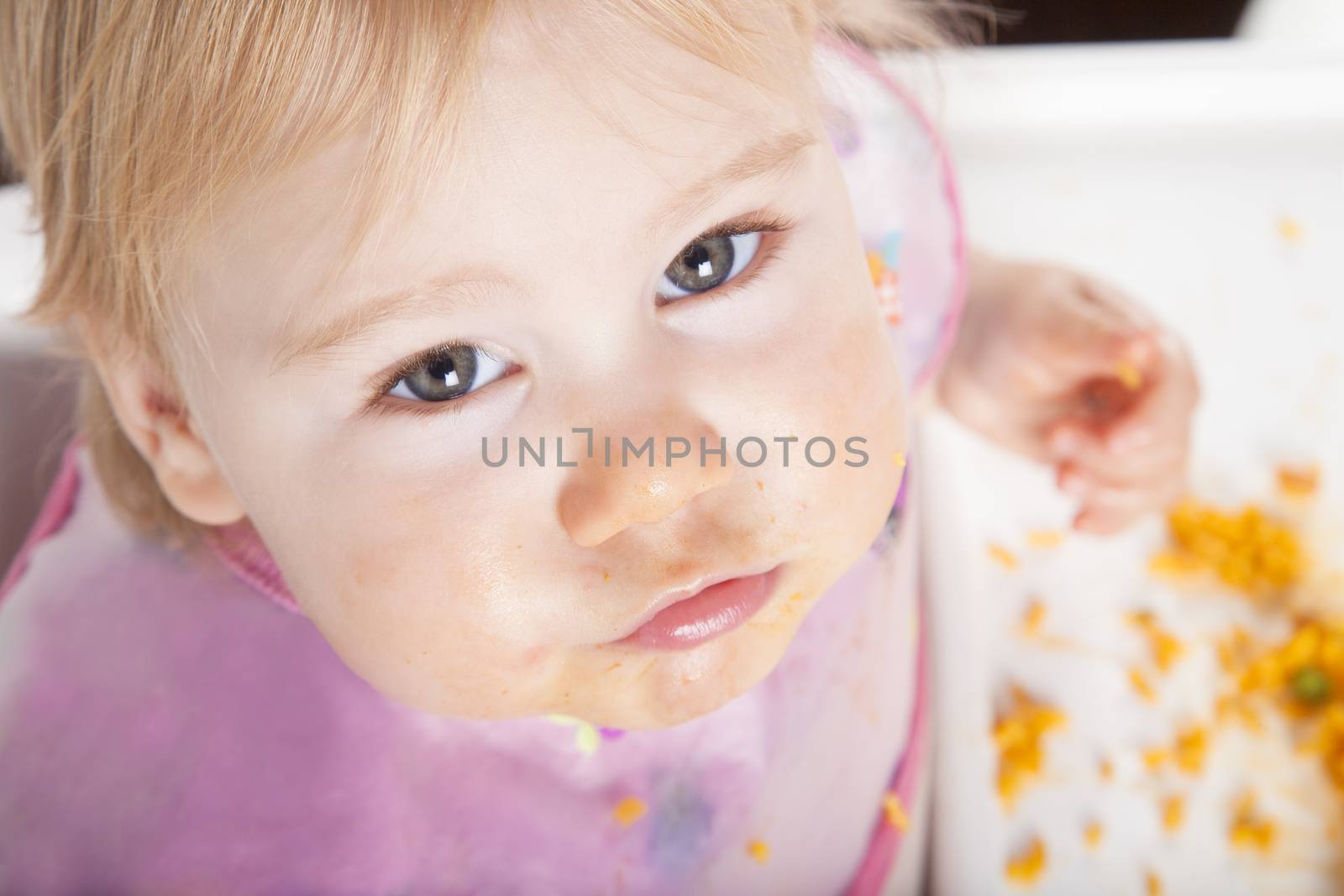 overhead shot of one year age caucasian baby pink plastic bib eating meal yellow orange rice paella with her hand in white high-chair
