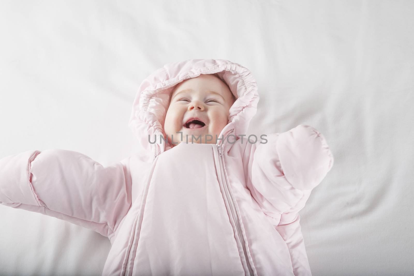 six months age cute baby dressed up in pink fluffy winter snowsuit snug hoodie clothes lying on white sheet bed smiling happy face