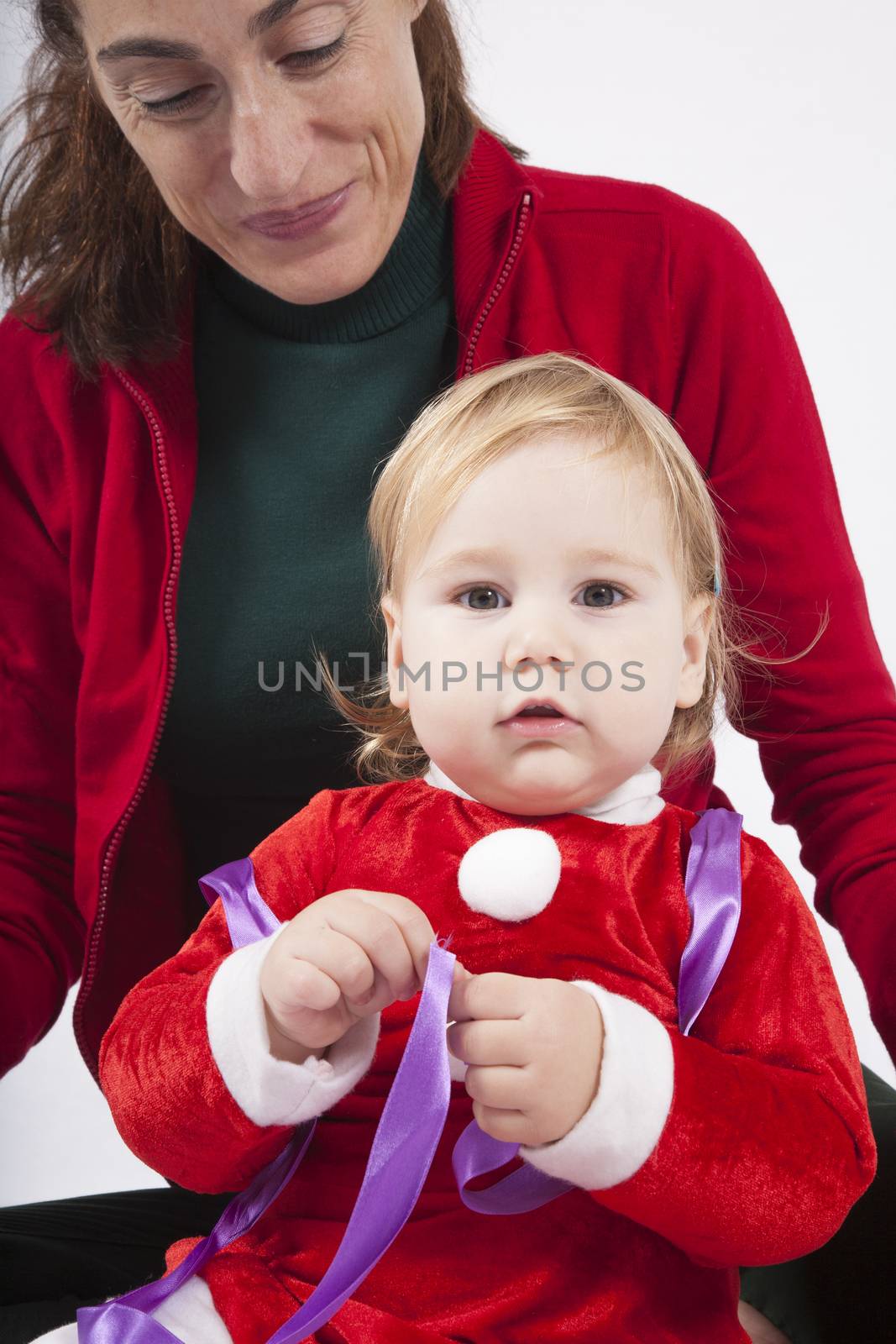 one year age caucasian blonde baby Santa Claus disguise with brunette woman mother red cardigan green trousers opening silver wrapped paper box gift Christmas on white background