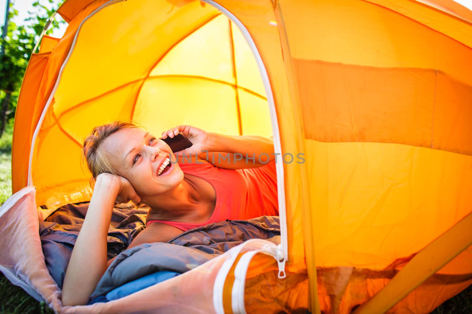 Pretty, young woman camping outdoors, lying in the tent in the morning, using her smart phone