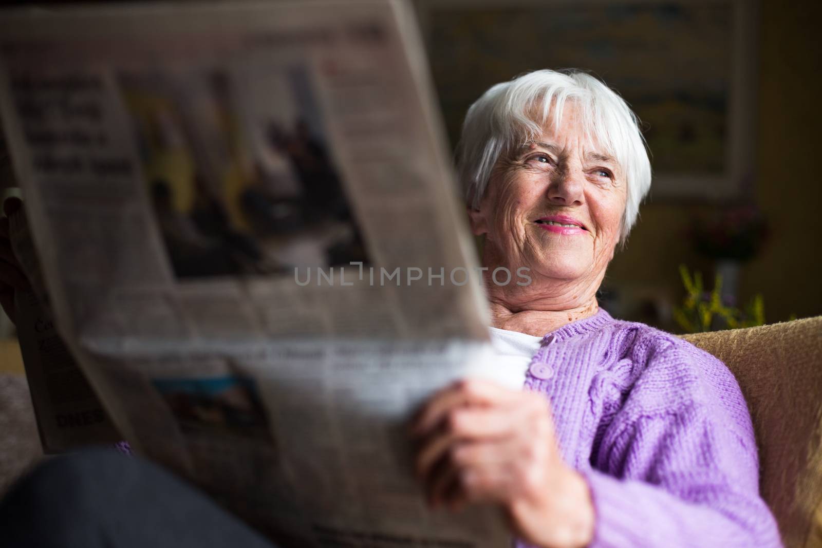 Senior woman reading morning newspaper, sitting in her favorite chair in her living room, looking happy