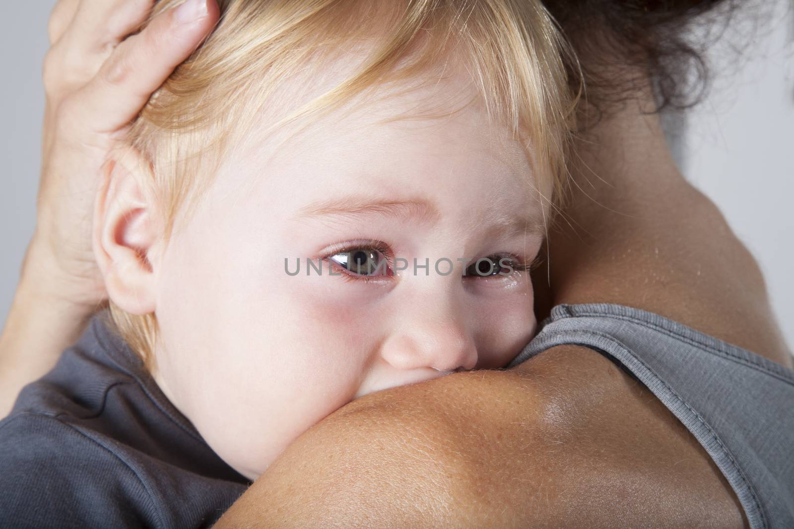 portrait of one year age blonde lovely cute caucasian white baby grey shirt looking  face crying and scream shout with tears eyes in brunette woman mother arms embrace hug