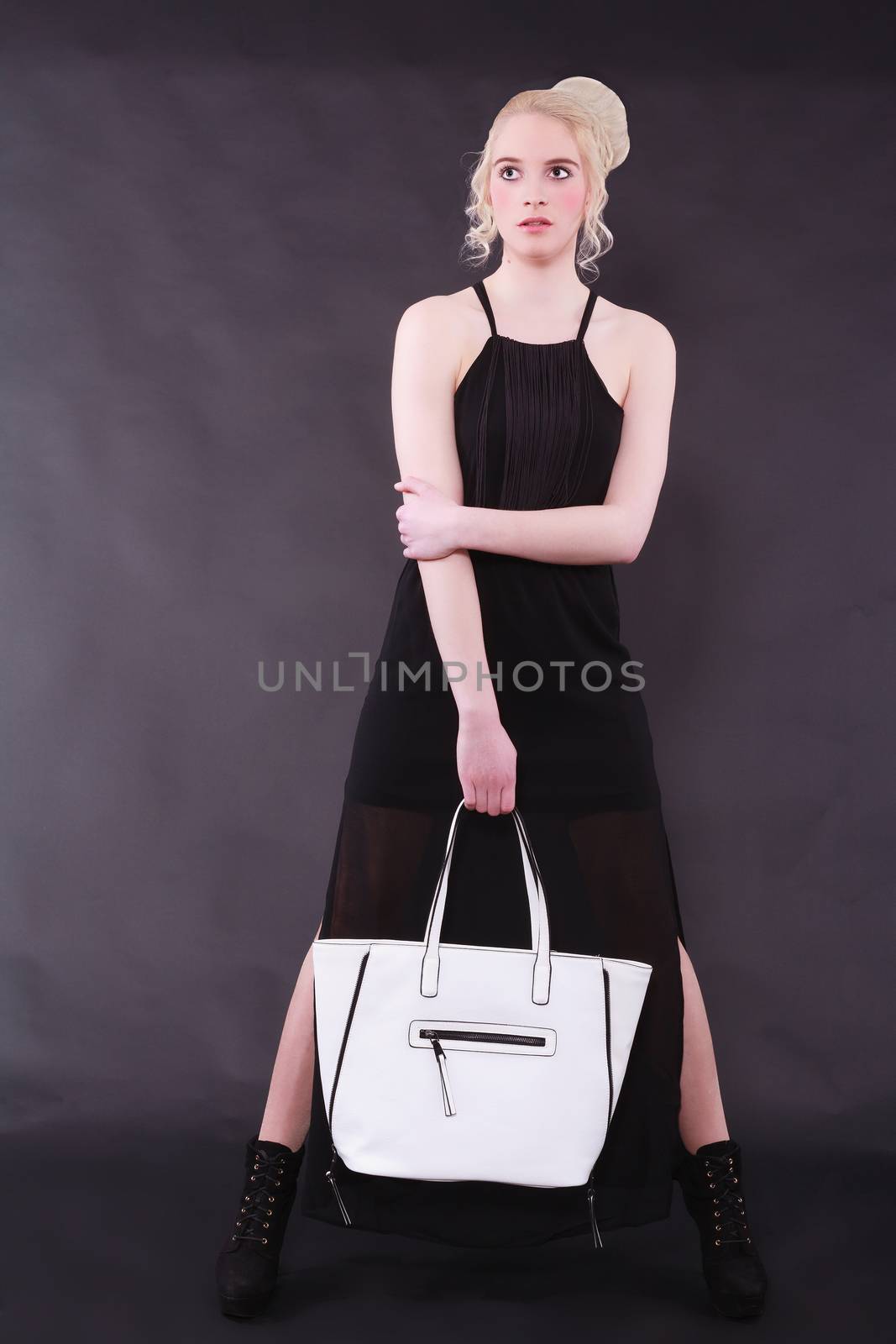 Blonde young model with modern hairstyle stands legs in black dress against a black background and holding a white bag in his hand.