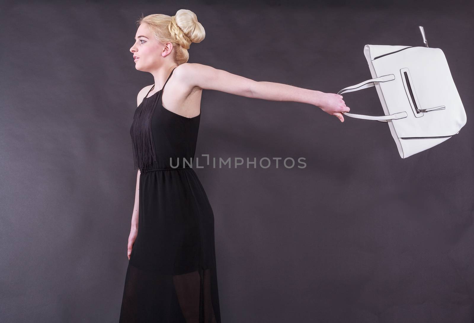 Lively young blond woman in a black dress with a white handbag on an outstretched arm back before black background, studio shot.