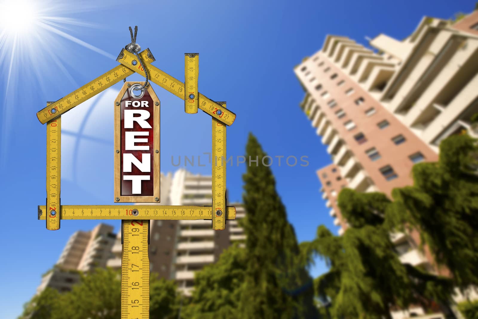 Yellow wooden meter ruler in the shape of house and label with text for rent. For rent real estate sign with tall and blurred buildings in the background