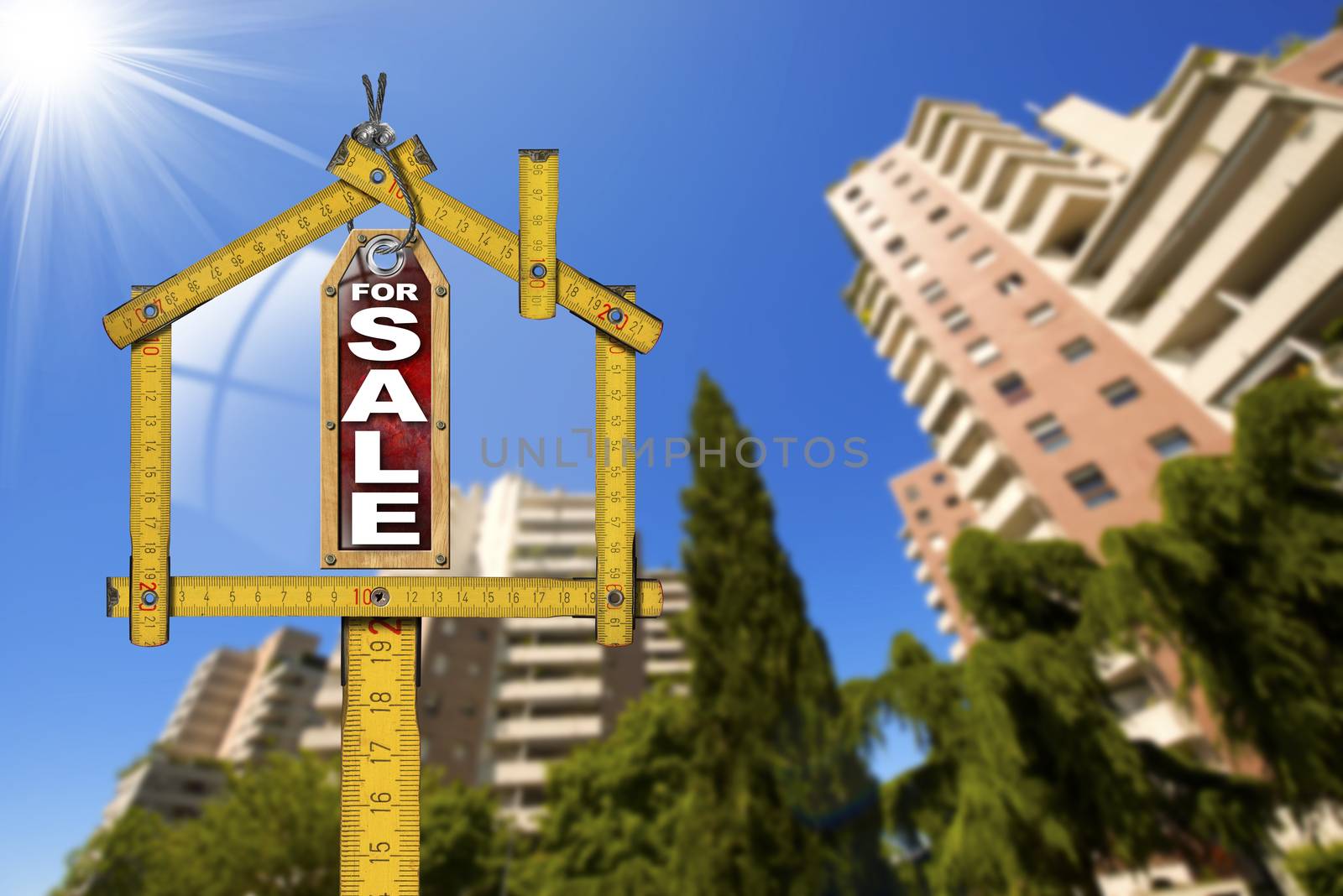 Yellow wooden meter ruler in the shape of house and label with text for sale. For sale real estate sign with tall and blurred buildings in the background