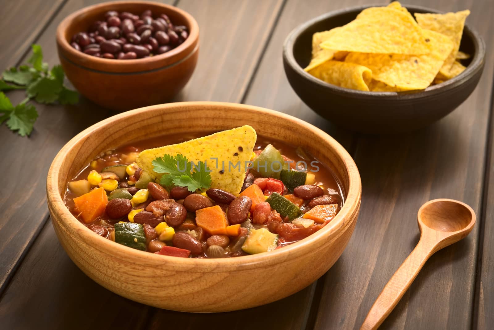 Wooden bowl of vegetarian chili dish made with kidney bean, carrot, zucchini, bell pepper, sweet corn, tomato, onion, garlic, garnished with tortilla chips and fresh coriander leaf, photographed with natural light (Selective Focus, Focus on the garnish)   