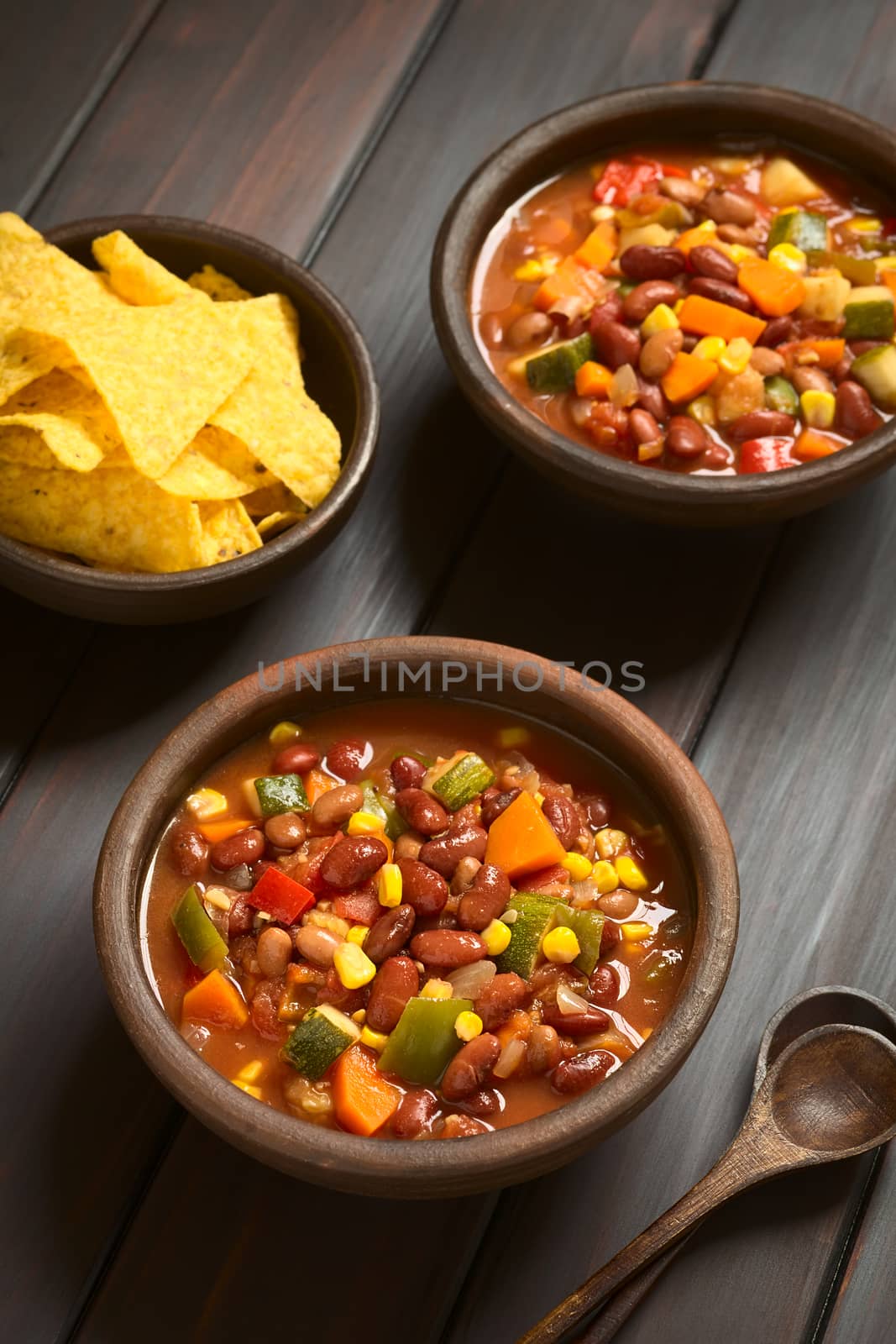 Two rustic bowls of vegetarian chili dish made with kidney bean, carrot, zucchini, bell pepper, sweet corn, tomato, onion, garlic, with tortilla chips on the side, photographed with natural light (Selective Focus, Focus in the middle of the first dish)   