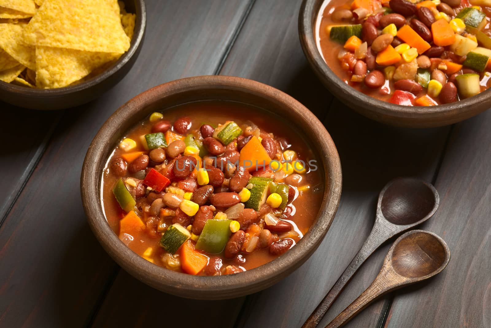 Two rustic bowls of vegetarian chili dish made with kidney bean, carrot, zucchini, bell pepper, sweet corn, tomato, onion, garlic, with tortilla chips on the side, photographed with natural light (Selective Focus, Focus in the middle of the first dish)   