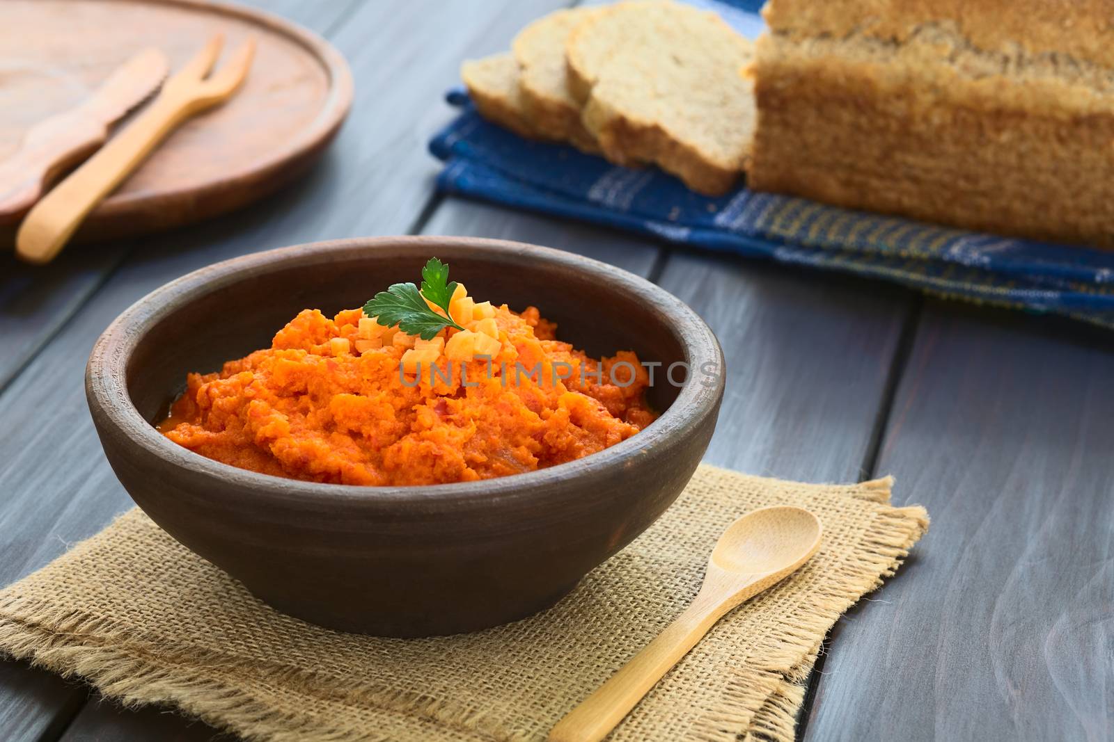 Carrot and red bell pepper spread in rustic bowl garnished with parsley leaf, wholegrain bread in the back, photographed with natural light (Selective Focus, Focus on the parsley leaf)