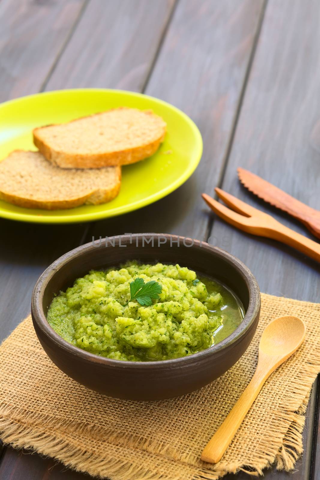 Rustic bowl of homemade zucchini and parsley spread garnished with fresh parsley leaf, slices of wholegrain bread in the back, photographed with natural light (Selective Focus, Focus on the leaf) 