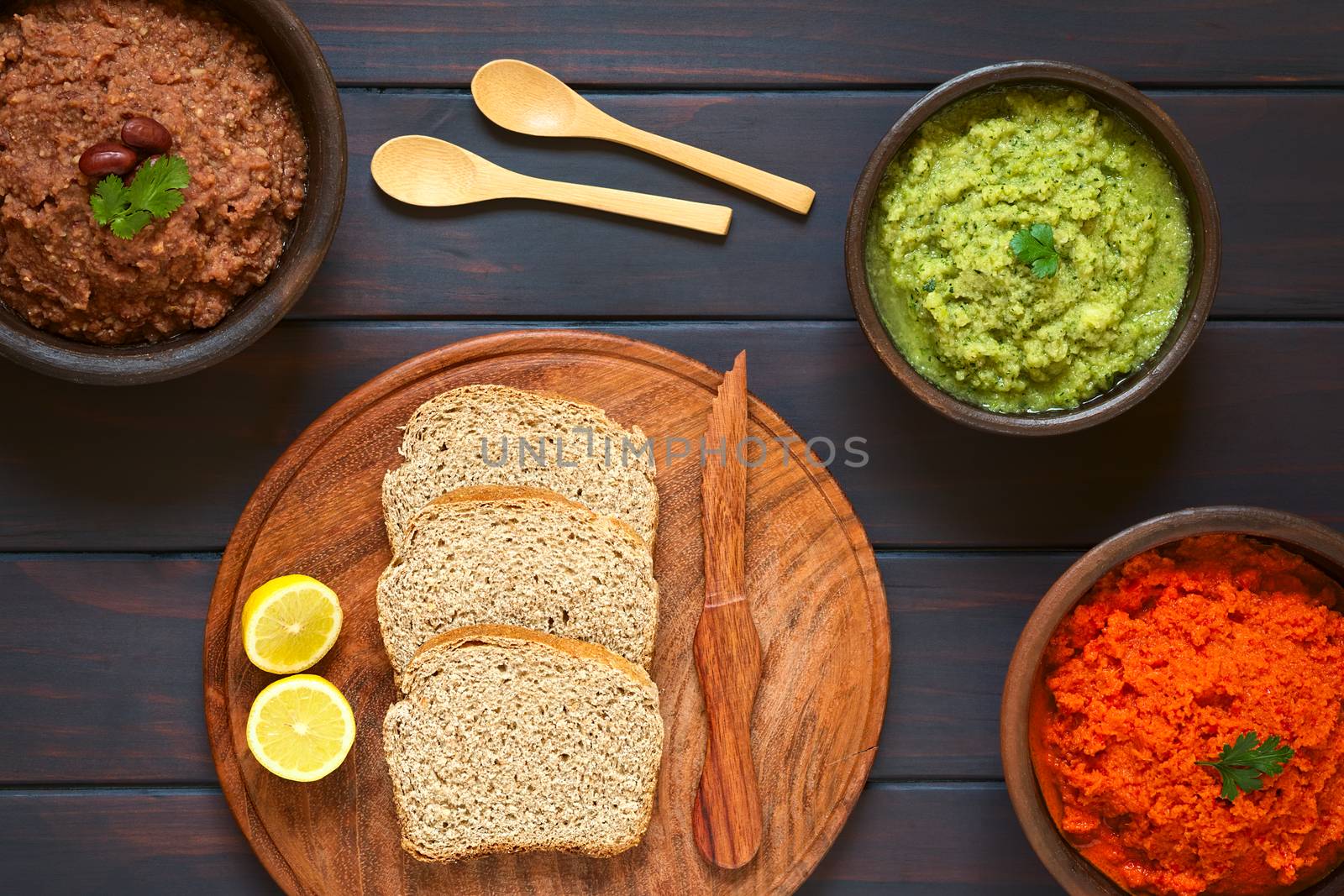 Overhead shot of wholegrain bread slices on wooden plate with three rustic bowls of homemade vegetable spreads (red kidney bean, zucchini and parsley, carrot and red bell pepper), photographed on dark wood with natural light