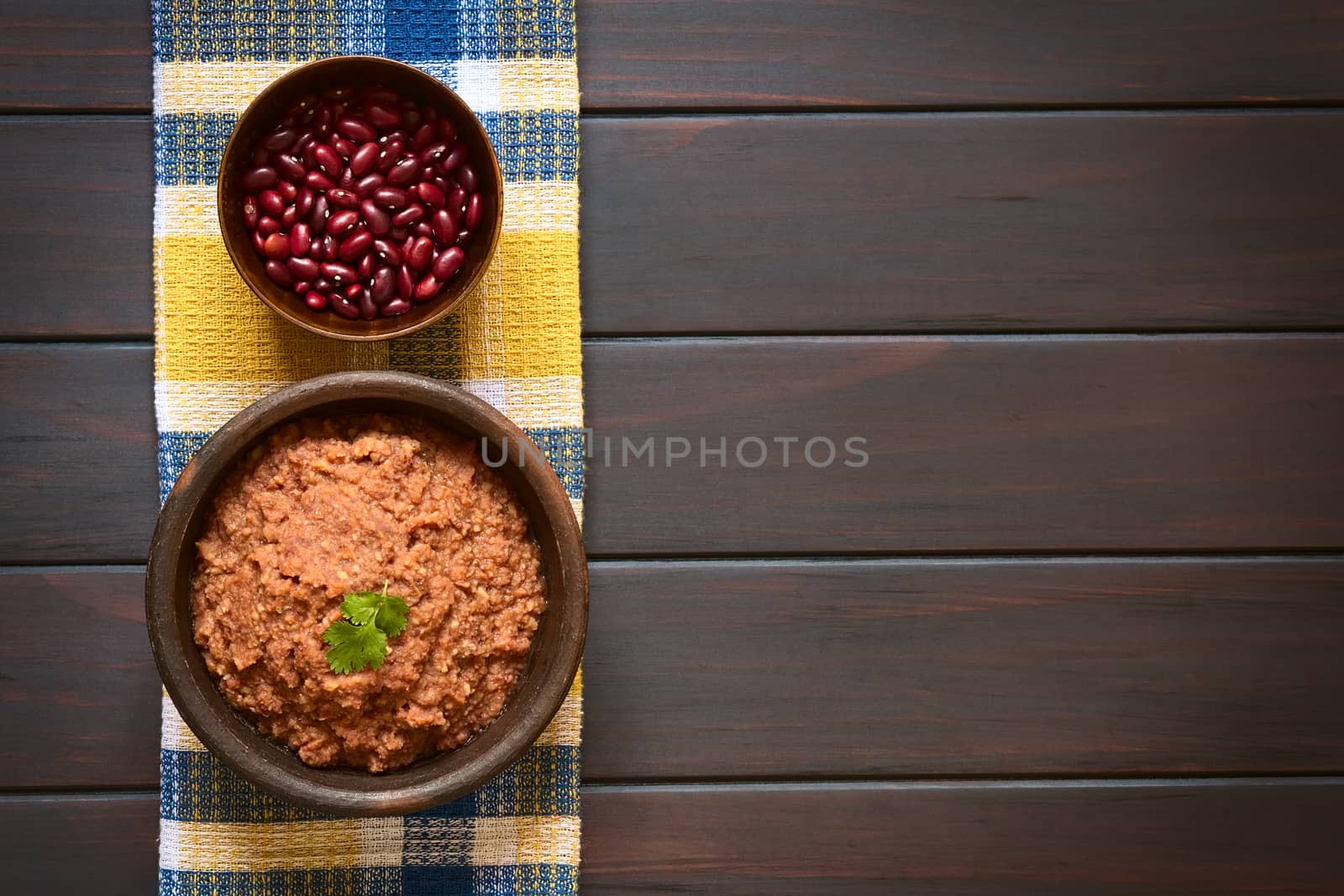 Overhead shot of a rustic bowl of homemade red kidney bean spread garnished with fresh coriander leaf, a bowl of raw kidney beans above, photographed on dark wood with natural light