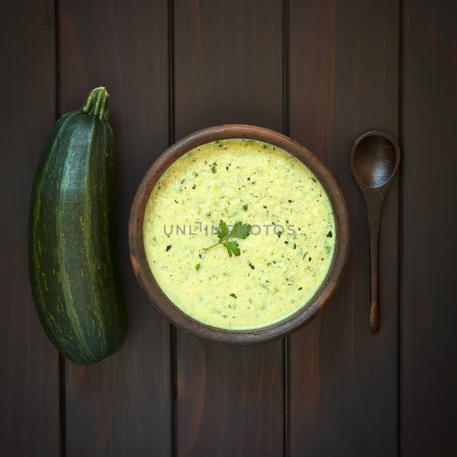 Overhead shot of a rustic bowl of cream of zucchini soup garnished with parsley leaf, spoon and zuchini on the side, photographed on dark wood with natural light