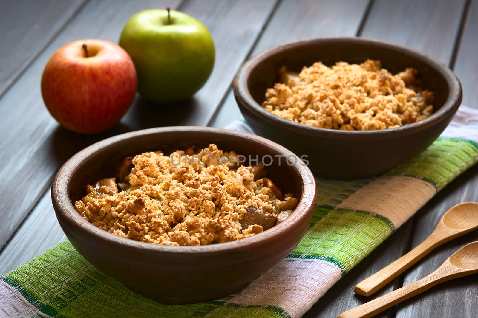 Two rustic bowls of baked apple crumble or crisp on kitchen towel, wooden spoons and fresh apples on the side, photographed on dark wood with natural light (Selective Focus, Focus one third into the first crumble)