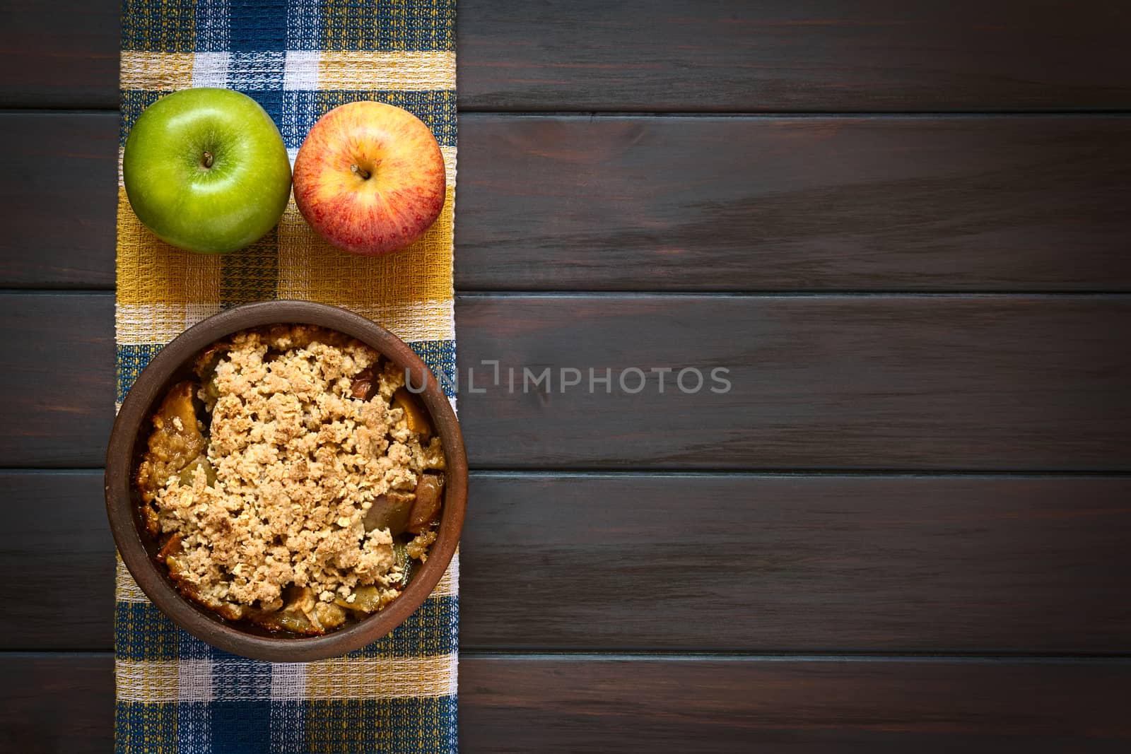 Overhead shot of a rustic bowl of baked apple crumble or crisp with fresh apples on a kitchen towel, photographed on dark wood with natural light
