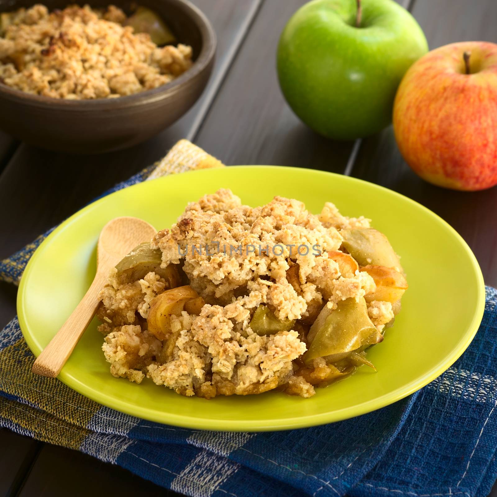 Freshly baked apple crumble or crisp served on plate with wooden spoon, fresh apples and a rustic bowl of apple crumble in the back, photographed on dark wood with natural light (Selective Focus, Focus one third into the crumble)
