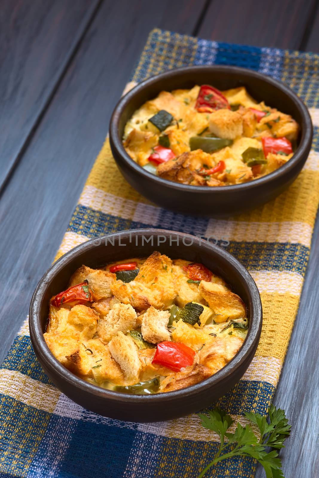 Savory baked vegetarian bread pudding made of zucchini, bell pepper, tomato and diced baguette, seasoned with thyme and parsley in rustic bowl on kitchen towel, photographed on dark wood with natural light (Selective Focus, Focus in the middle of the first dish)