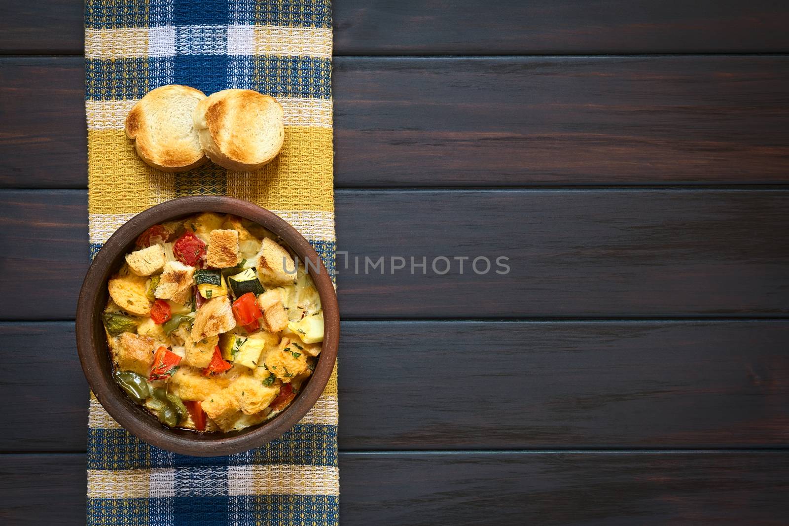Overhead shot of savory baked vegetarian bread pudding made of zucchini, bell pepper, tomato and diced baguette, seasoned with thyme and parsley in rustic bowl, with two toasted baguette slices on kitchen towel, photographed on dark wood with natural light