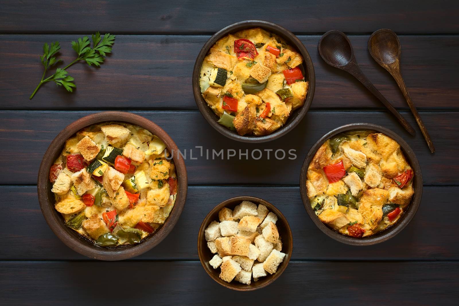 Overhead shot of savory baked vegetarian bread pudding made of zucchini, bell pepper, tomato and diced baguette, seasoned with thyme and parsley in rustic bowls, ingredients and wooden spoons on the side, photographed on dark wood with natural light