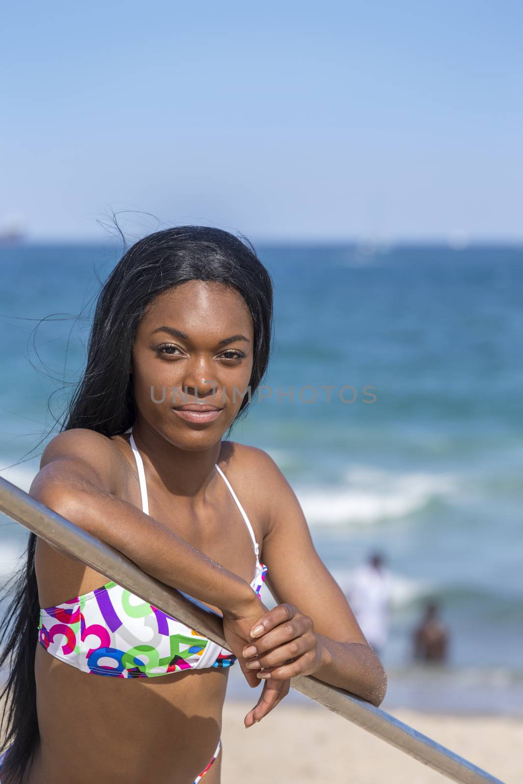 Young black woman at the beach, wearing a bikini. Leaning on railing. Blue sky.