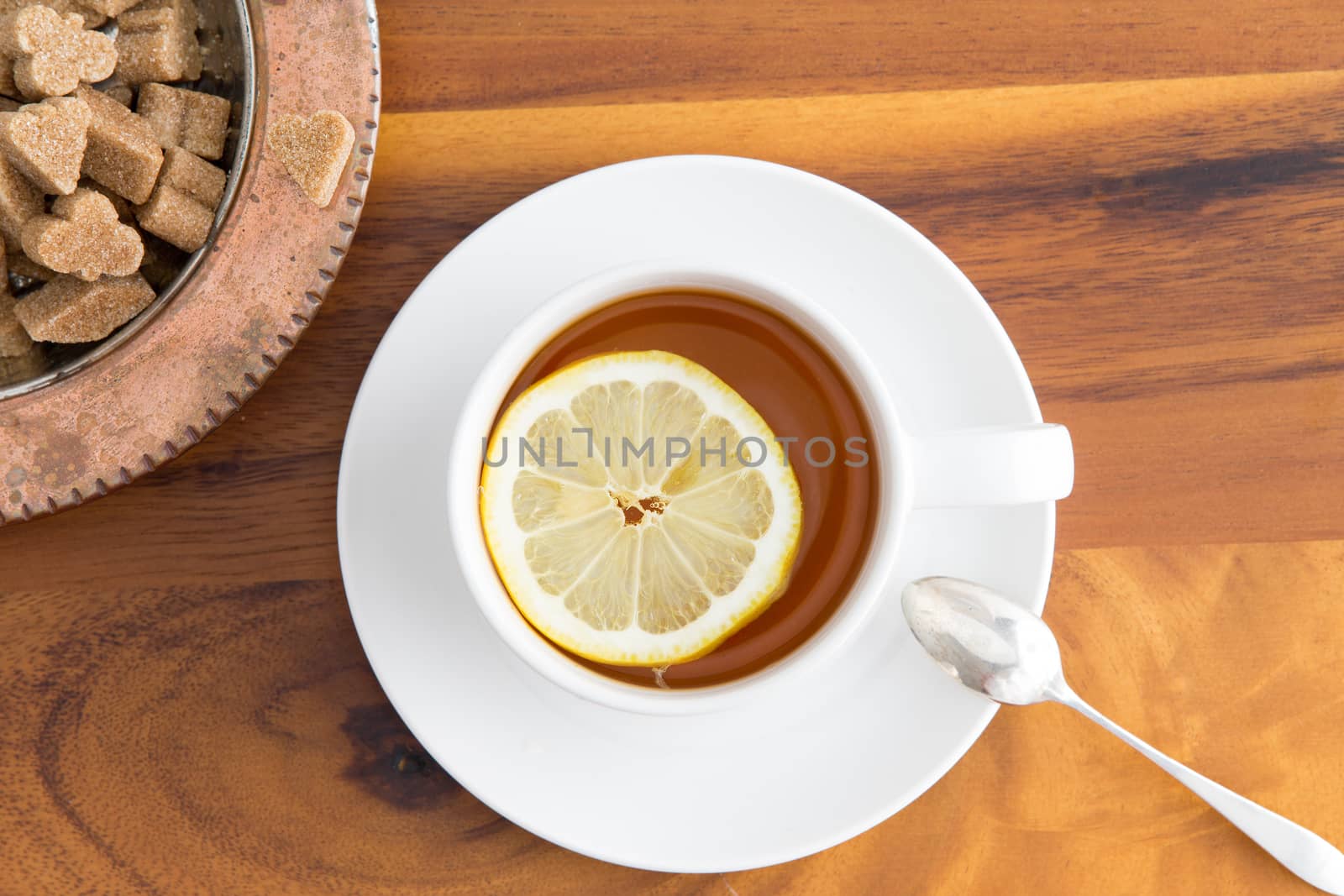 Cup of herbal tea served in a white cup and saucer with a slice of lemon and brown sugar cubes in a sugar bowl on a wooden table viewed from above