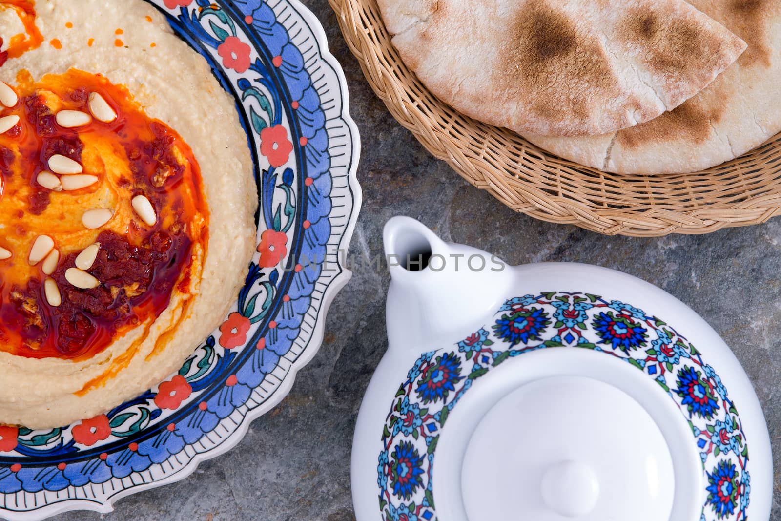 Bowl of savory nutritious hummus, a creamy dip of mashed chickpeas, tahini, oil, lemon juice, and garlic, served with pita bread to use as a scoop for the dip and a pot of tea, overhead view