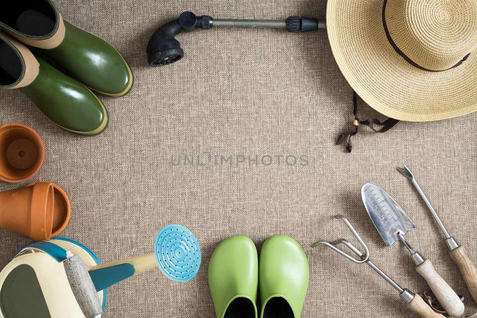 Border or frame of gardening tools on a neutral beige textile background with garden shoes and boots, sunhat, watering can, hand tools and flowerpots, with central copyspace, view from above