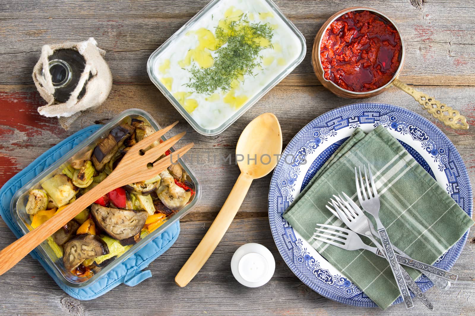 Grilled vegetables with eggplant, tzatziki, Turkish cacik with dill, and hot red chili pepper paste ready to be served with plates and cutlery on an old weathered wooden table