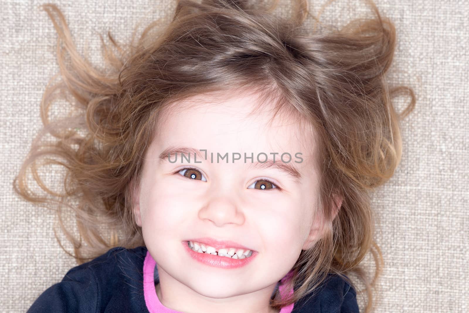 Headshot of young three year old girl lying on a carpet with tousled hair grinning up at the camera with a happy expression
