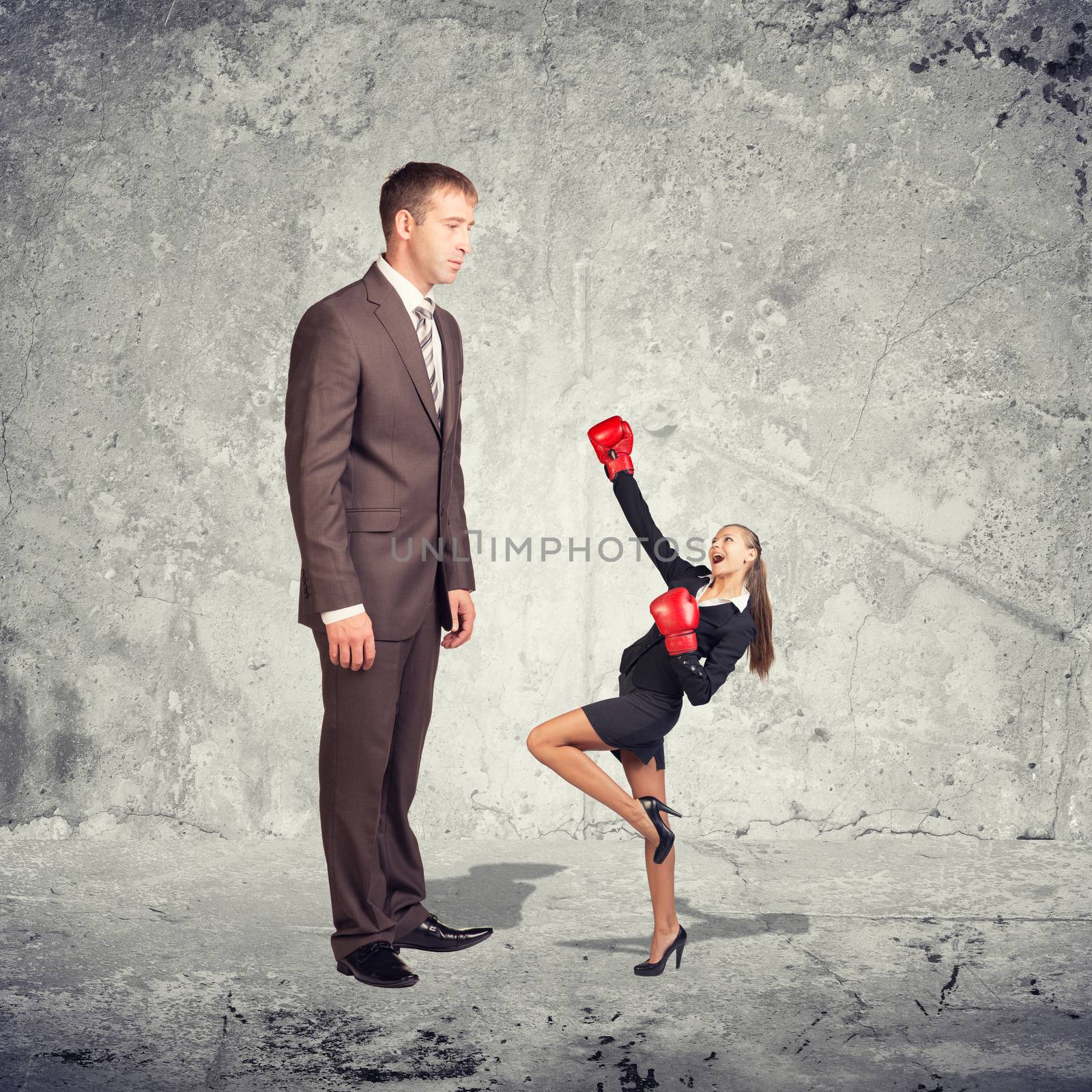 Large businessman looking at small businesswoman in boxing gloves. Concrete wall as backdrop by cherezoff
