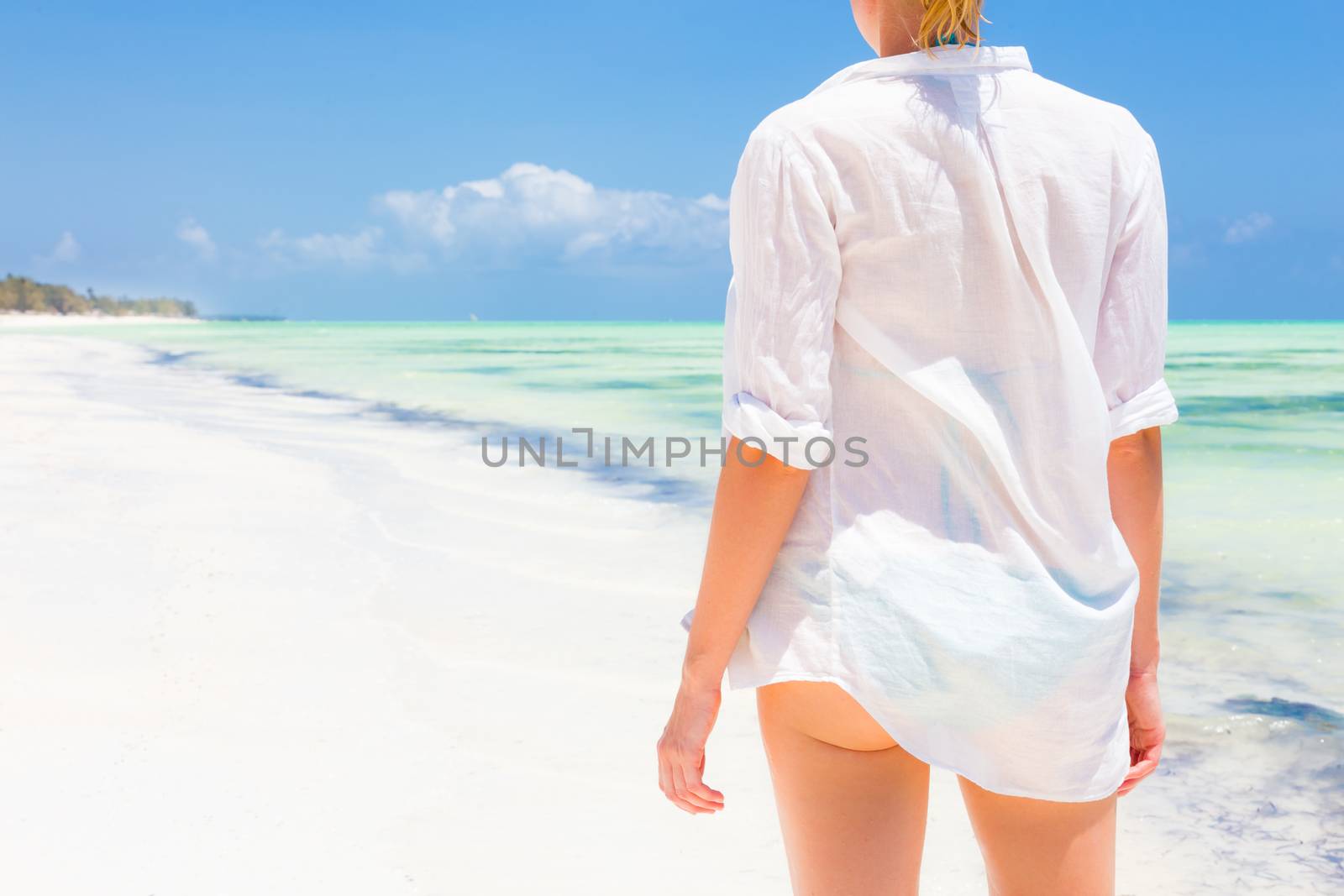 Happy woman enjoying, relaxing joyfully in summer by tropical blue water. Beautiful caucasian model  wearing white beach tunic on vacations looking down picture perfect Paje beach, Zanzibar, Tanzania.