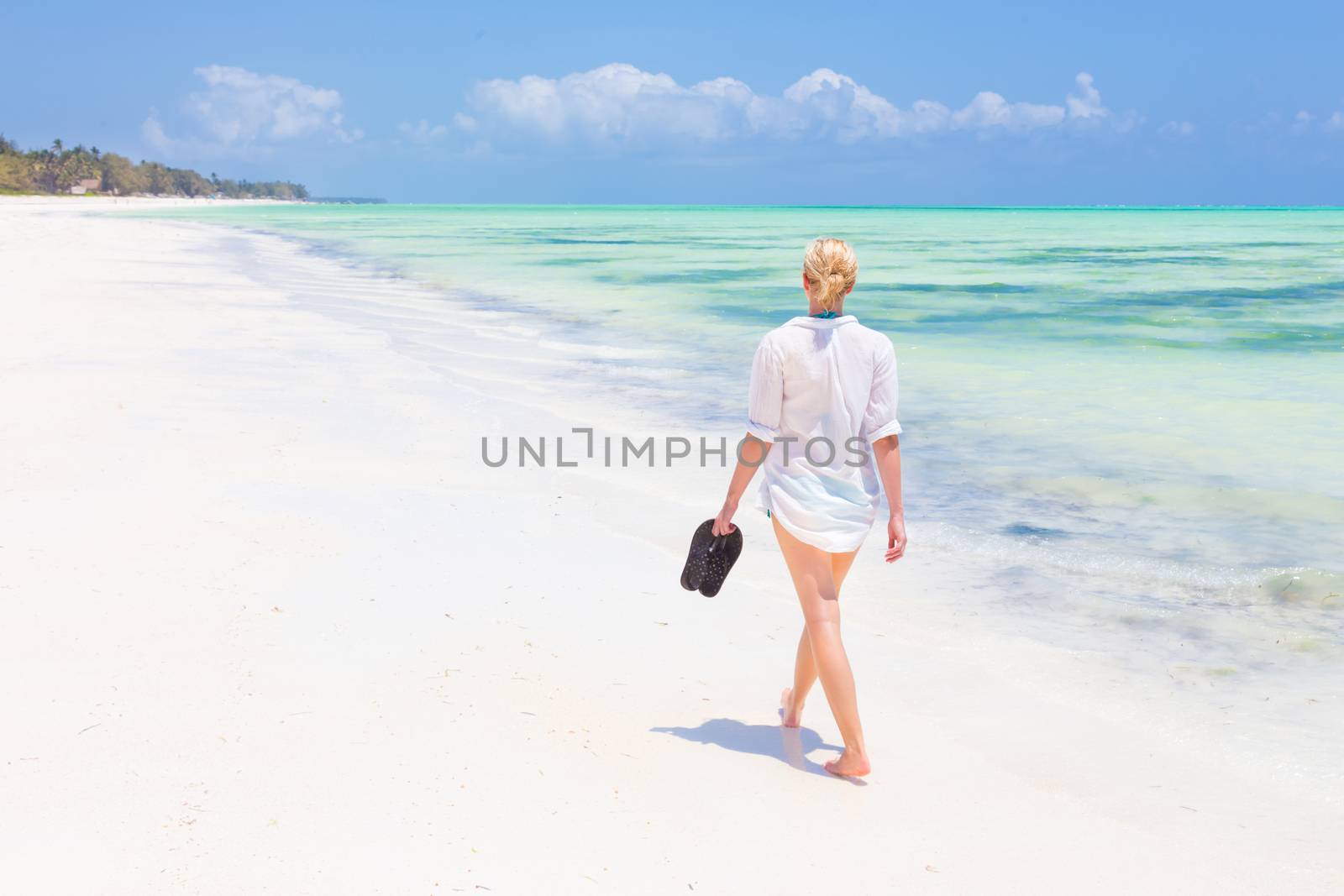 Caucasian woman walking joyfully on tropical beach. Beautiful caucasian model  wearing white beach tunic on vacations walking down picture perfect Paje beach, Zanzibar, Tanzania. Copy space.