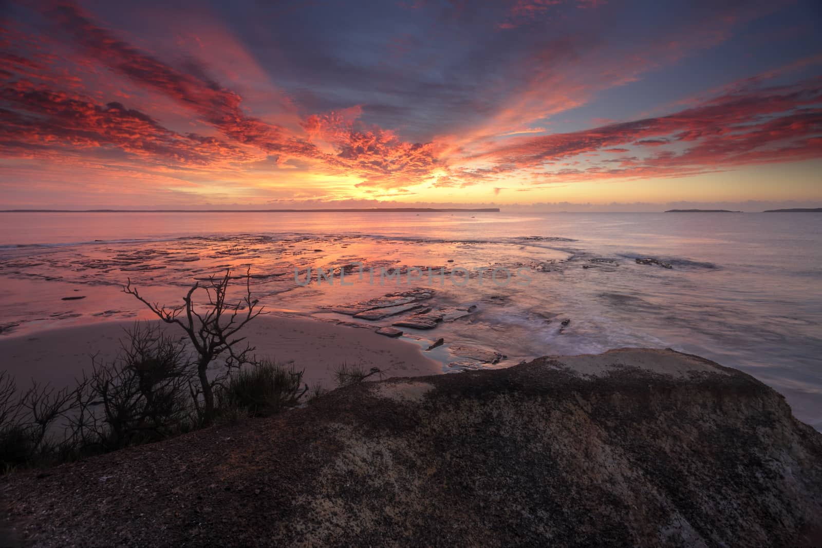 Spectacular sunrise over Plantation Point Vincentia.  Views from a rock ledge overlooking the reef at a high tide  and beyond to Beecroft Peninsula and Bowen Island on the horizon.  The brilliant colours in the sky also reflecting into the bay